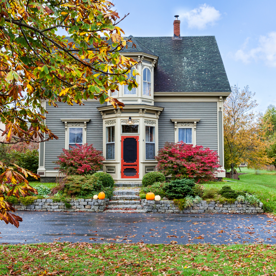 A grey house in Vermont with a red door and fall themed landscaping outside.