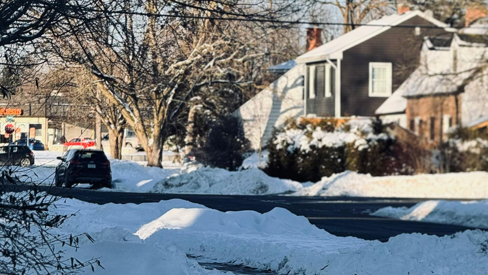 Snowy residential street with snowbanks and houses. A car drives on the road.