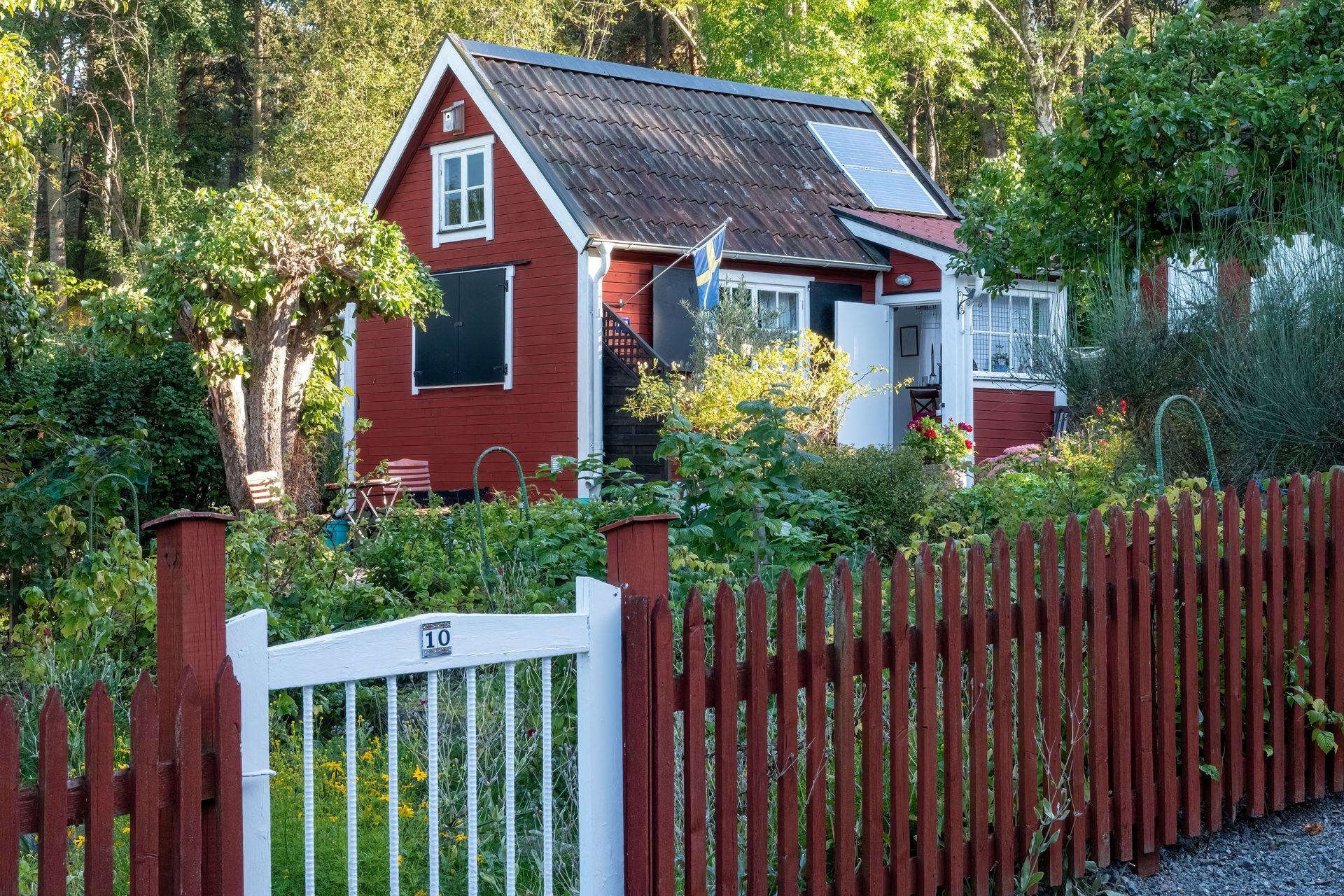 Small red cottage with white trim, a garden, and a red picket fence surrounded by trees.