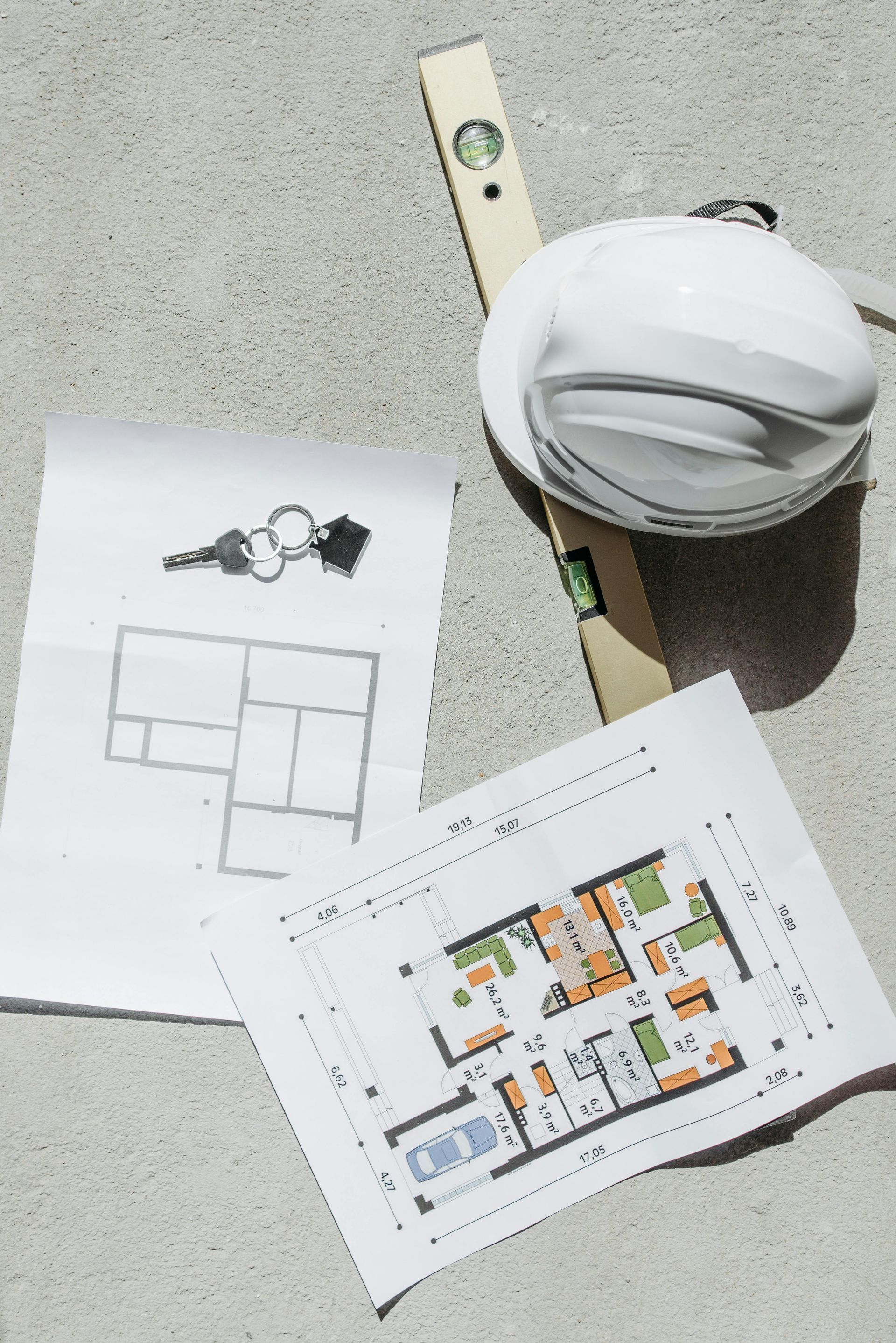 Architectural floor plans, a set of keys, a construction level, and a hard hat resting on a textured concrete surface.