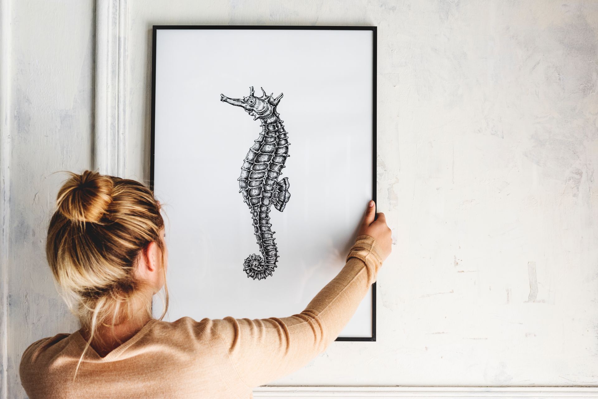A blonde woman hanging a hand drawing of a seahorse on the wall