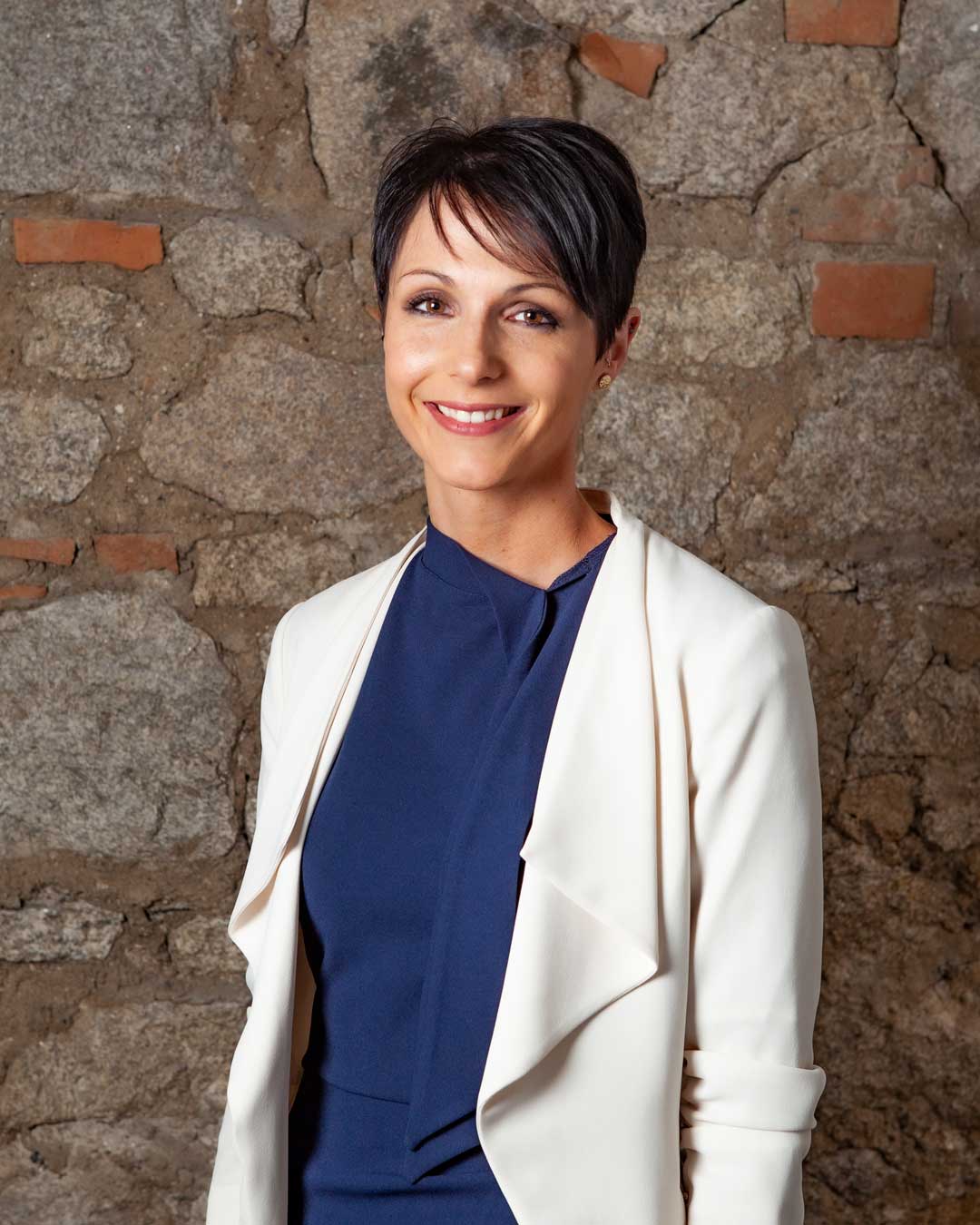 A woman in a blue dress and white jacket is smiling in front of a stone wall.