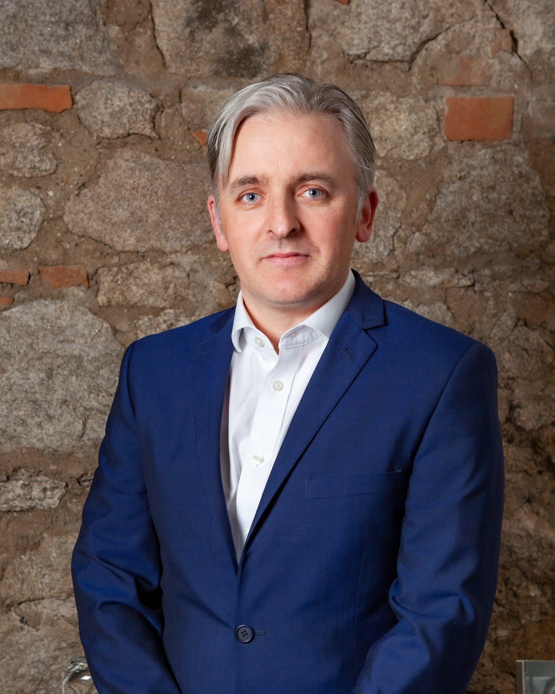 A man in a blue suit and white shirt is standing in front of a stone wall.