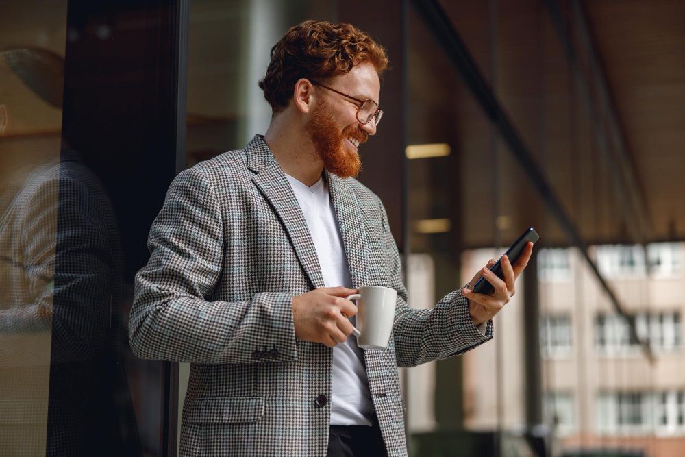 A man with a beard is holding a cup of coffee and looking at his cell phone.