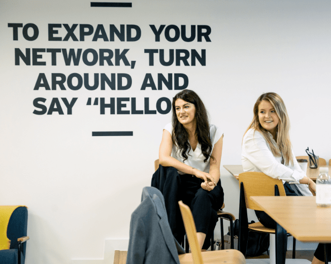 Two women sit in front of a wall that says to expand your network turn around and say hello