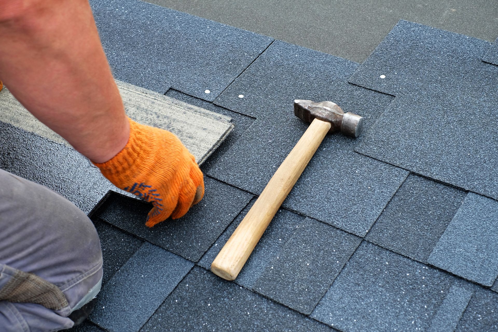 Roofing contractor hands with orange gloves, installing bitumen roof shingles using hammer in nails.