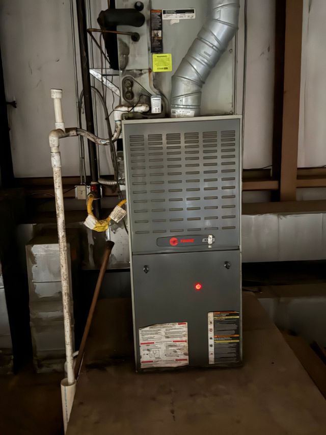 Basement furnace with silver ductwork, pipes, and a red indicator light on the front panel