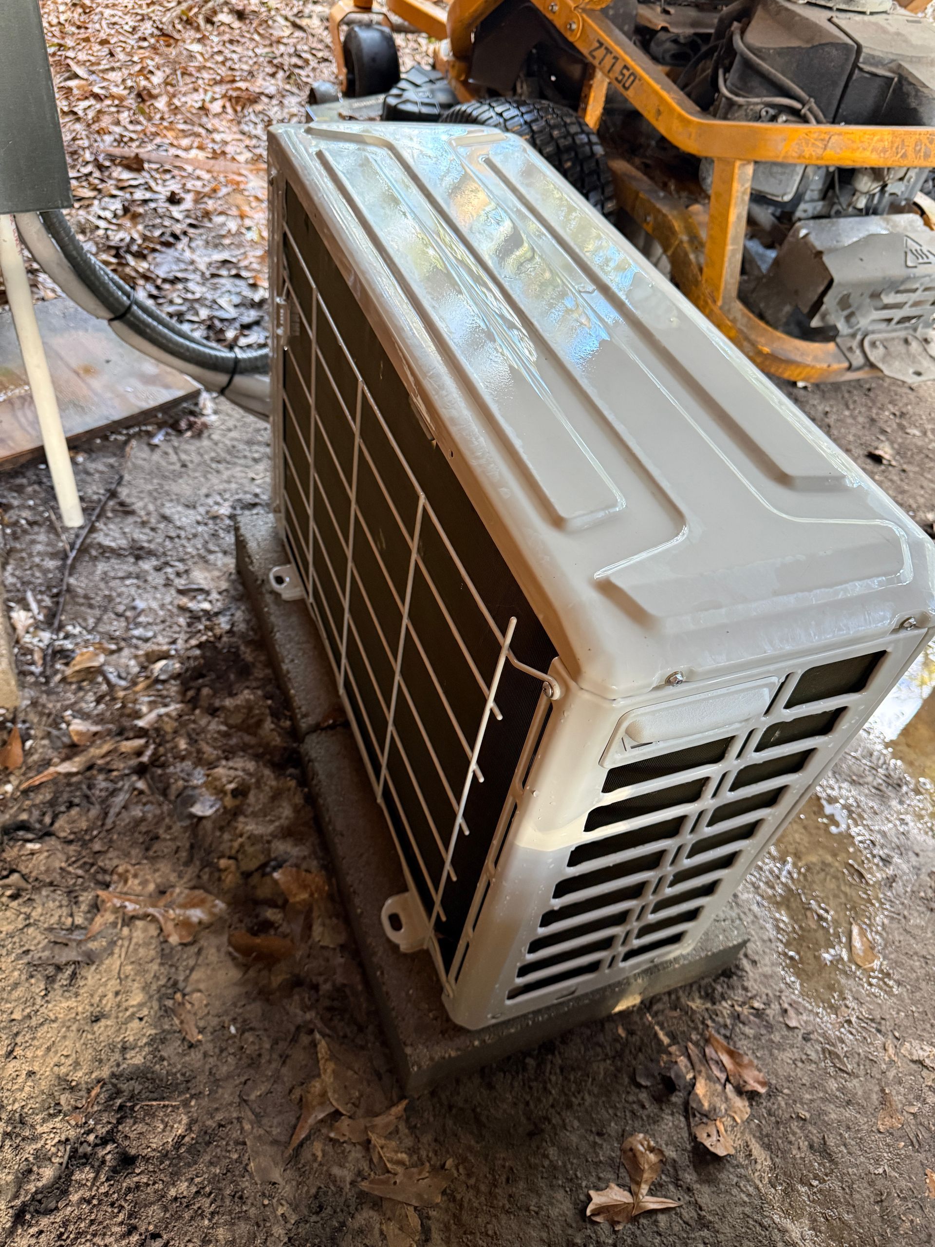 Outdoor air conditioner unit lying on its side in a muddy work area next to a yellow machine.