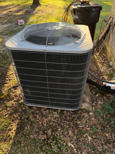 Outdoor air conditioner unit on grass beside a house, with a trash bin in the background.