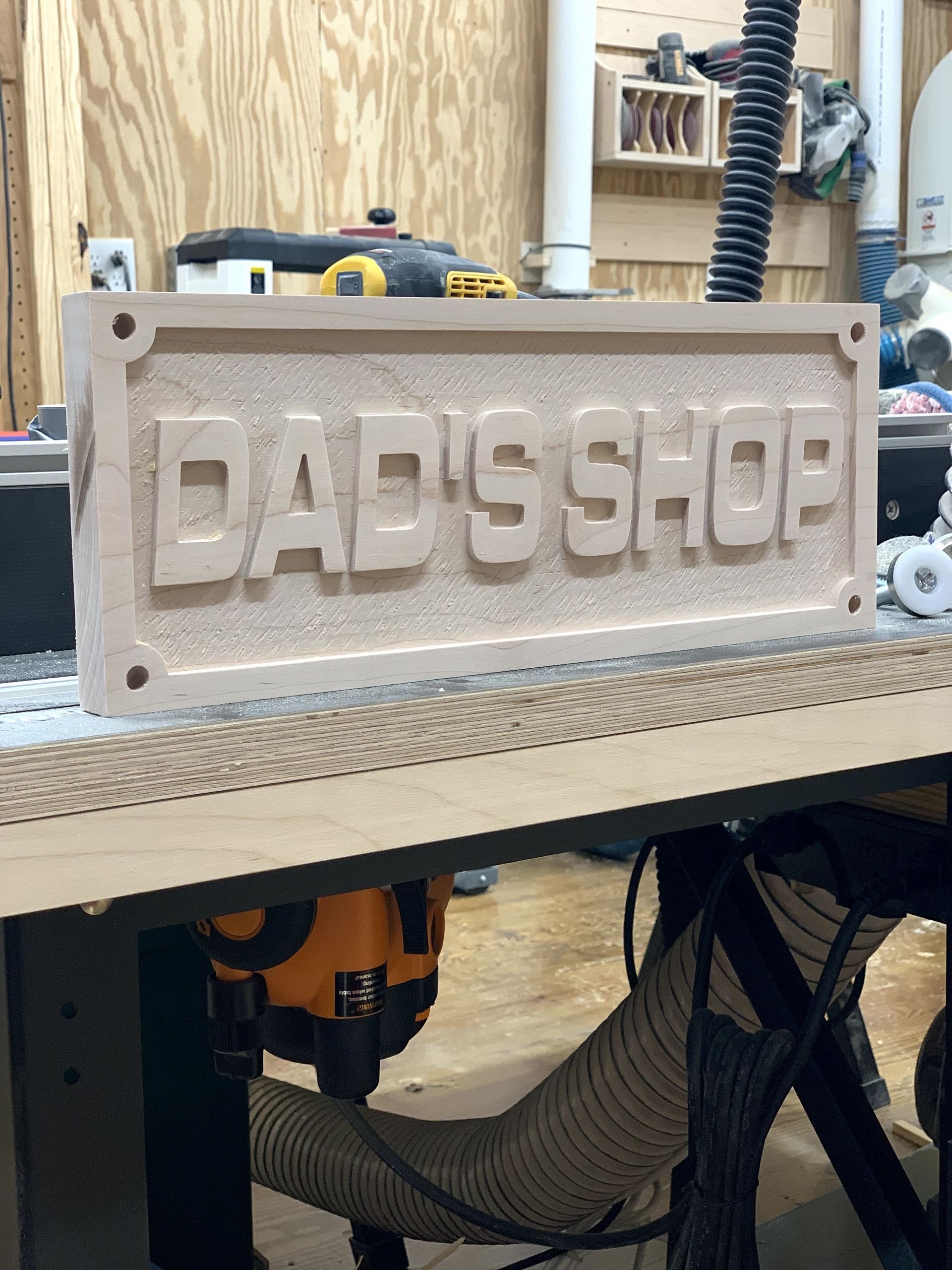 A wooden sign that says dad 's shop is being carved on a machine.