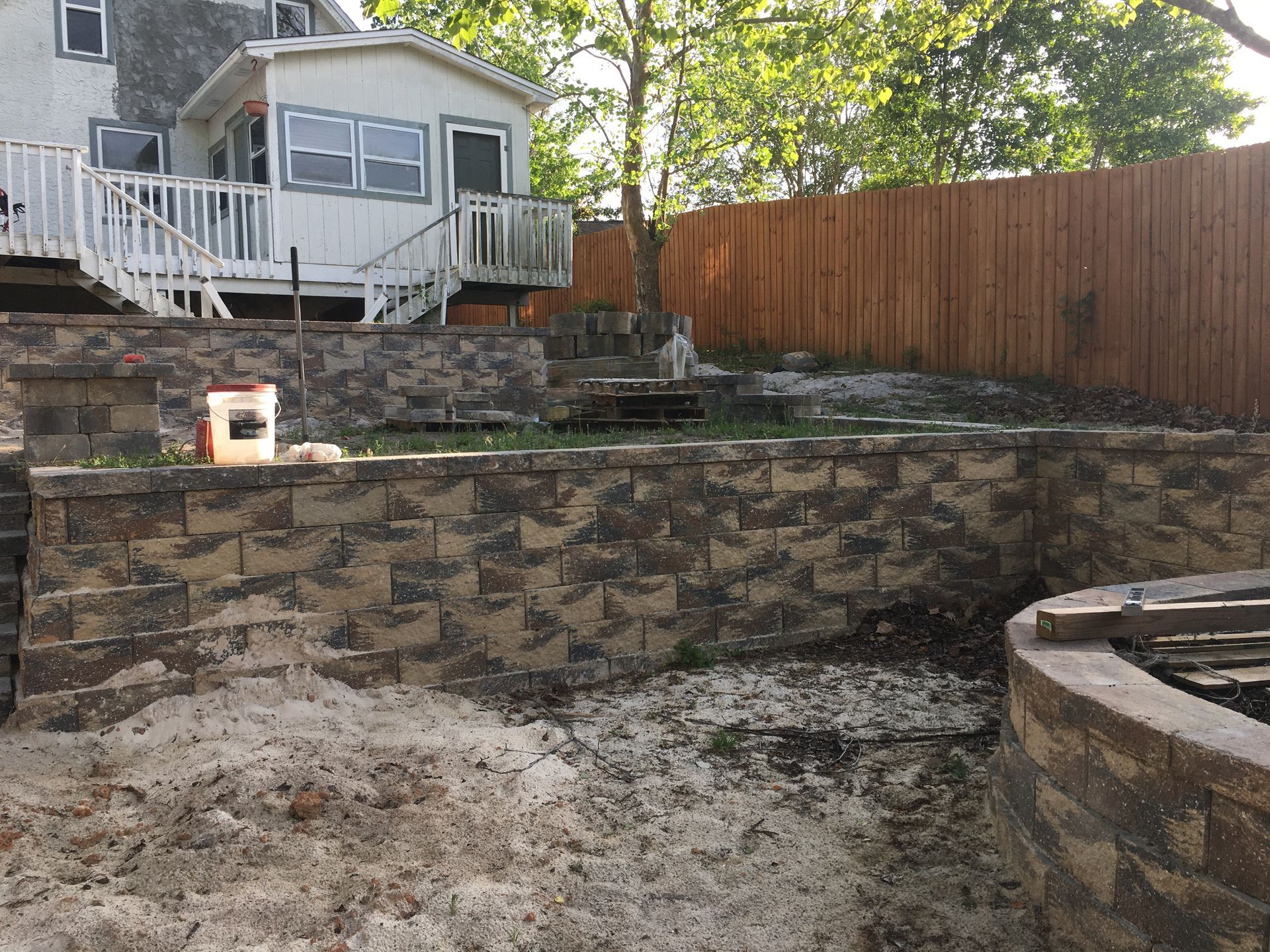A backyard with a stone wall and a wooden fence and a house in the background.