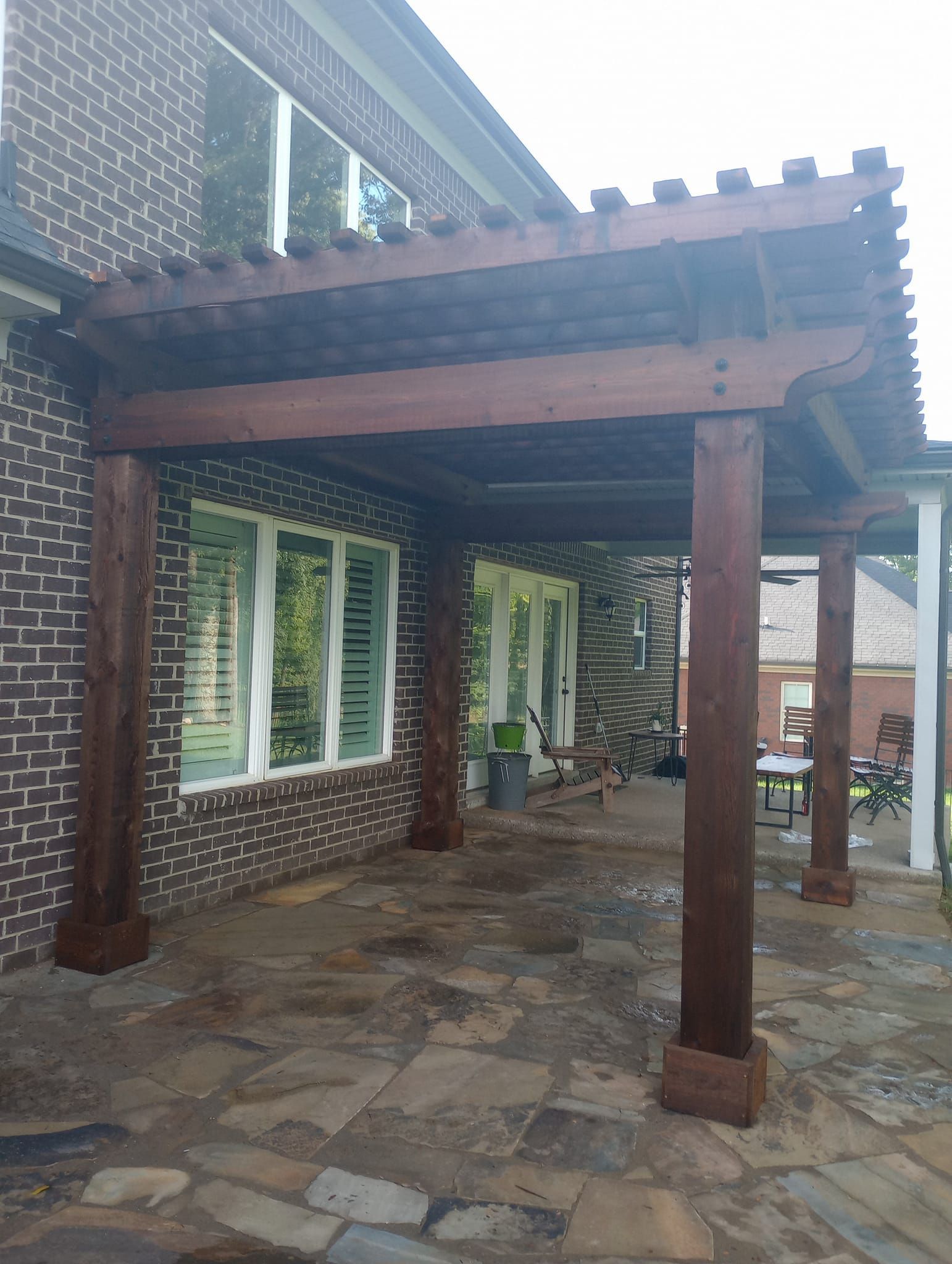 A wooden pergola is sitting on top of a stone patio in front of a brick building.