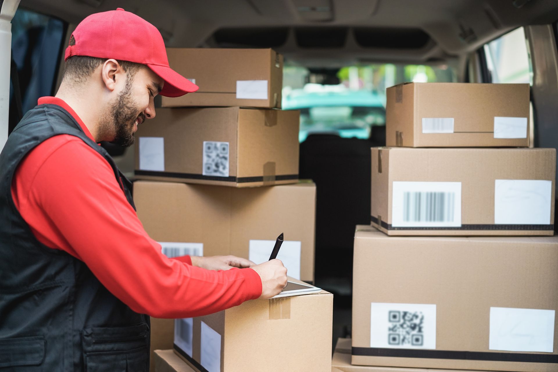A courier in a red shirt and cap smiles while writing on a cardboard box inside a delivery van filled with packages.