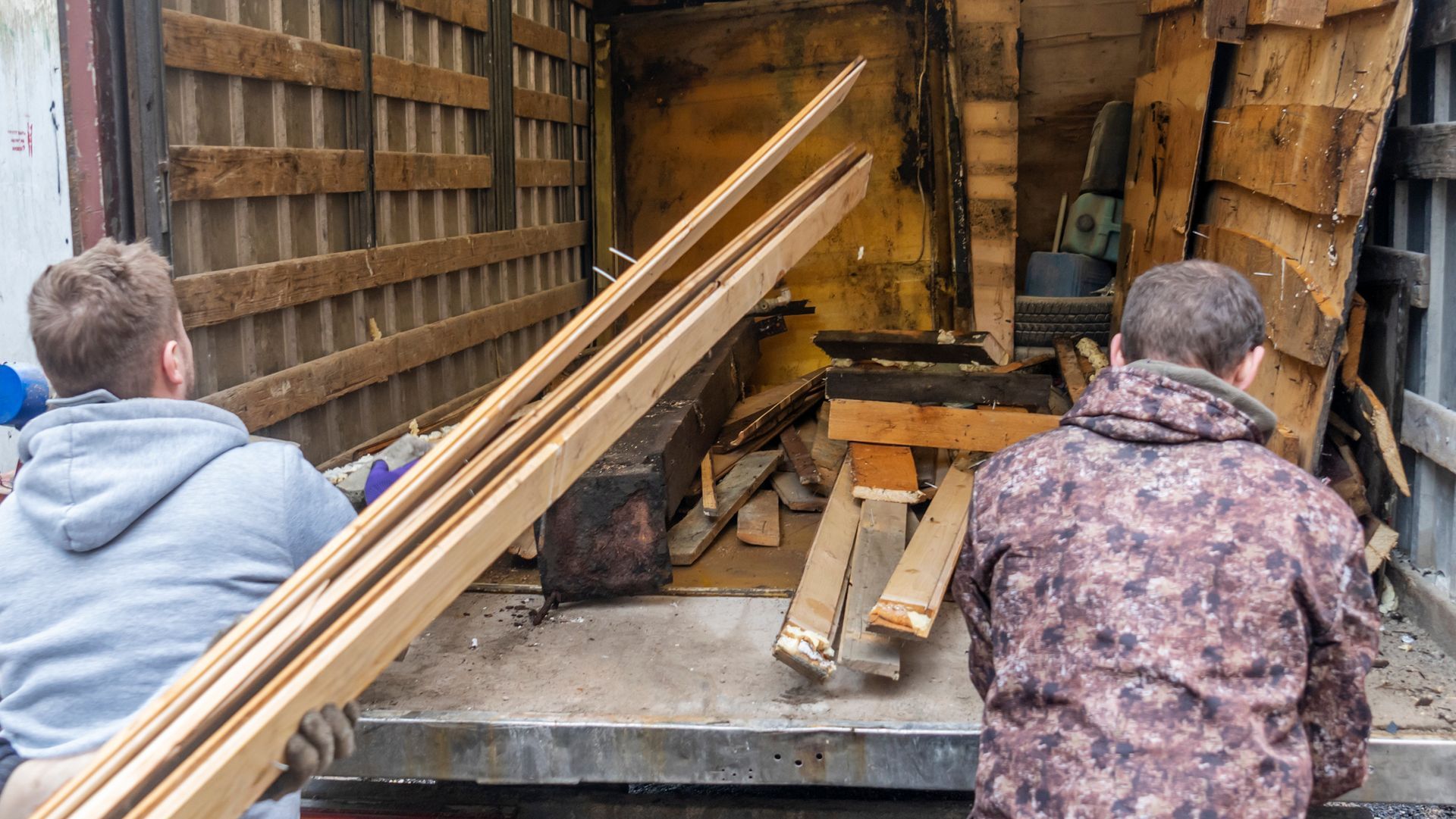 Two people load wooden planks into the back of a truck, surrounded by scattered timber and construction debris.