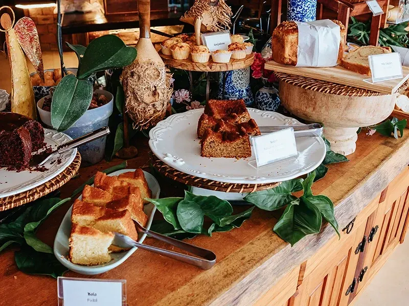 Uma mesa de buffet de madeira exibe diversos bolos, pães e doces em pratos brancos e suportes elevados de madeira.