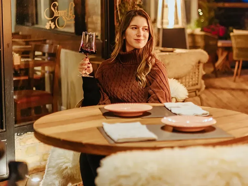 Uma pessoa vestindo um suéter marrom está sentada a uma mesa redonda de madeira, segurando uma taça de vinho tinto, em um restaurante ao ar livre.