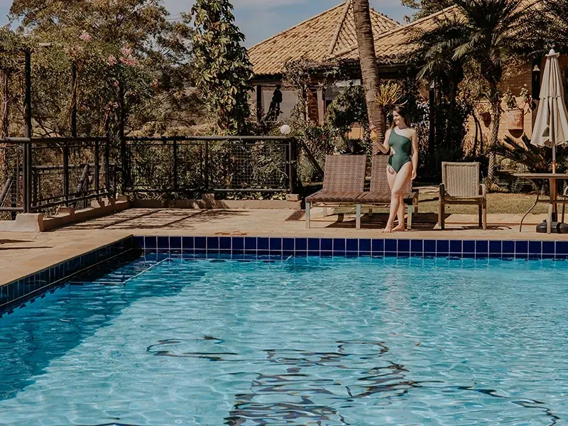 Uma pessoa vestindo um maiô verde-escuro está em pé ao lado de uma piscina azul-clara em um dia ensolarado, perto de espreguiçadeiras.