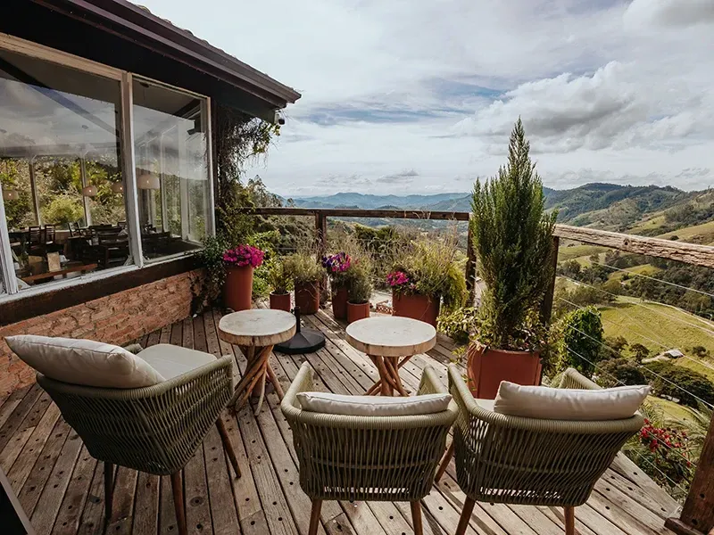 Três poltronas de vime em uma varanda de madeira com vista para uma paisagem montanhosa, com mesinhas e vasos de plantas.