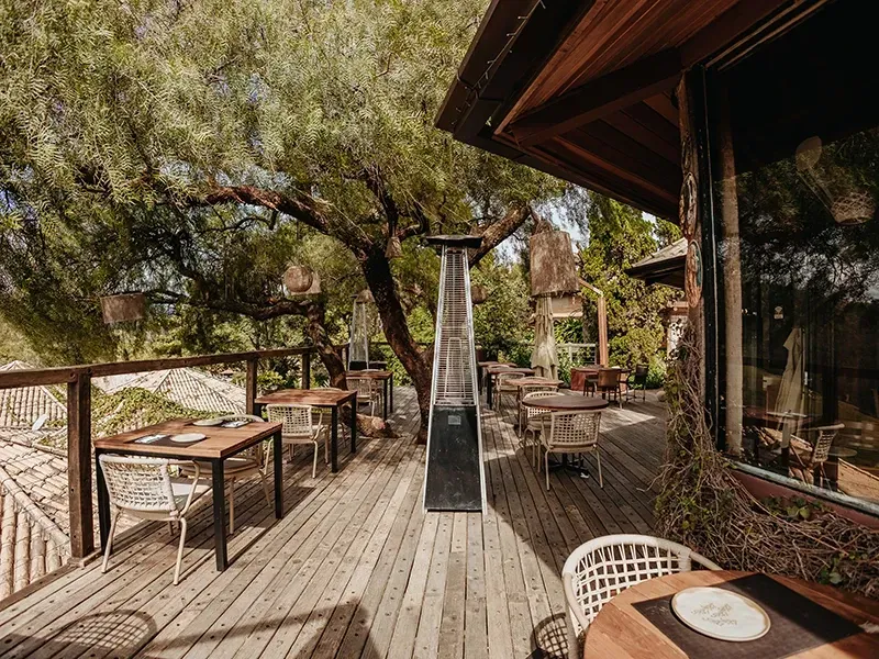 Terraço de restaurante ao ar livre com mesas e cadeiras de madeira em um deck, sombreado por árvores, com um aquecedor alto no centro.