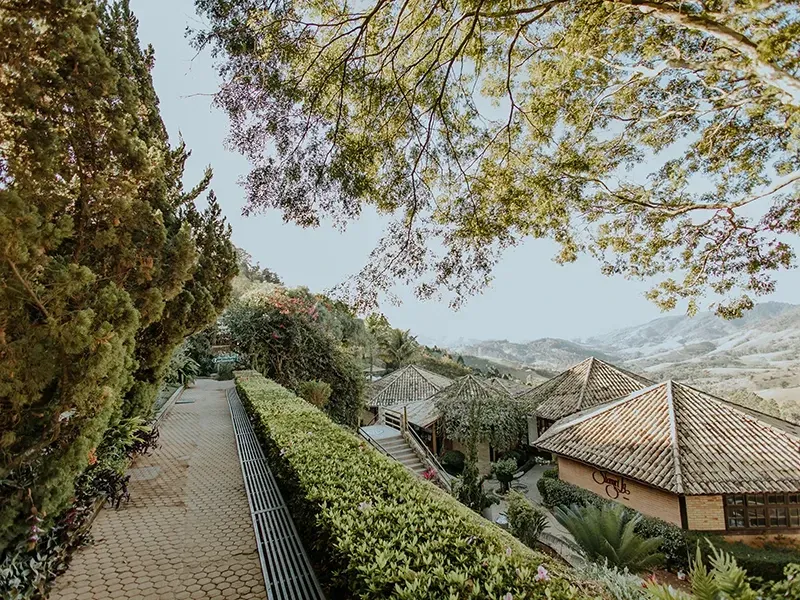Um caminho de pedra ladeado por árvores altas e sebes conduz a edifícios com telhados de telha, tendo como pano de fundo uma paisagem montanhosa deslumbrante.