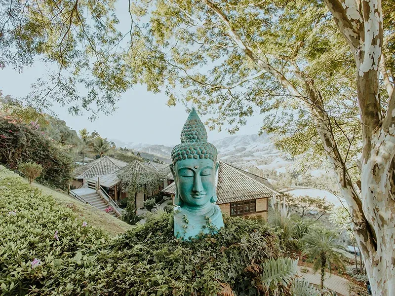 Uma escultura serena da cabeça de Buda, em tons de turquesa, está situada no centro de um exuberante jardim verde na encosta de uma colina, com vista para as montanhas.