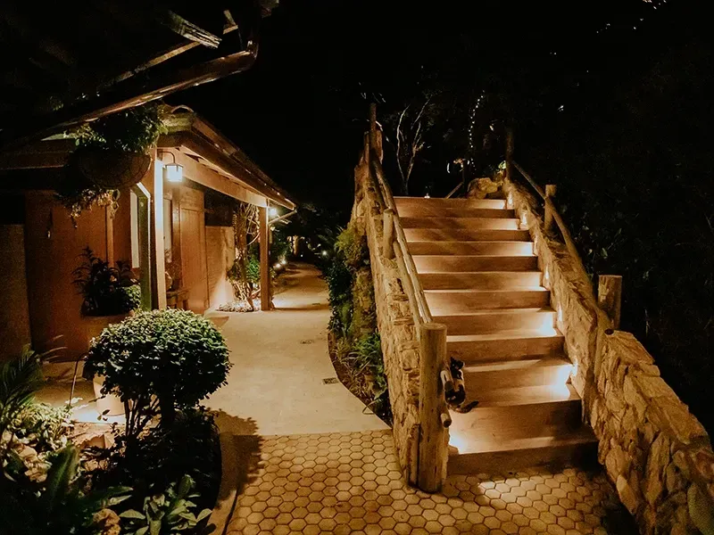Vista noturna de uma escadaria de pedra iluminada por luzes quentes, ao lado de um edifício e um caminho de jardim paisagístico.
