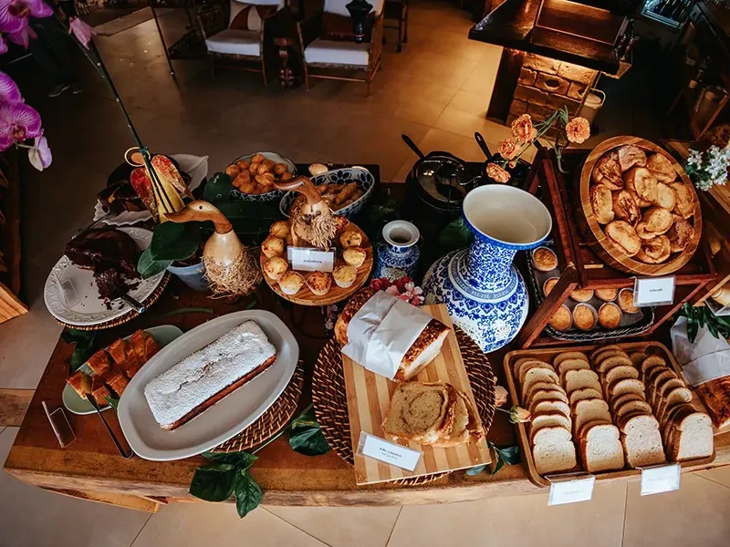Uma mesa de buffet de café da manhã de madeira repleta de diversos tipos de pães, doces, um bolo e peças decorativas de cerâmica azul e branca.