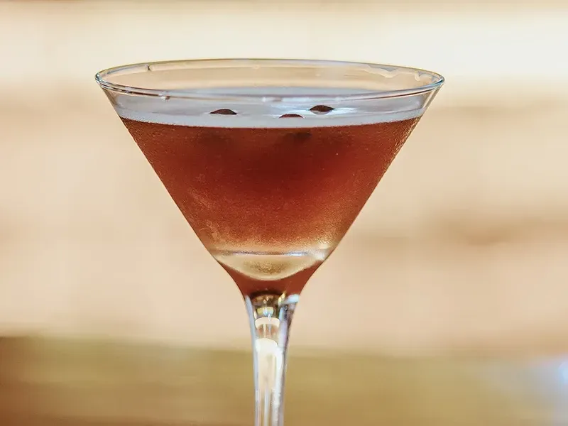 Um coquetel marrom-avermelhado com três grãos de café na superfície, servido em uma taça de martini contra um fundo desfocado.