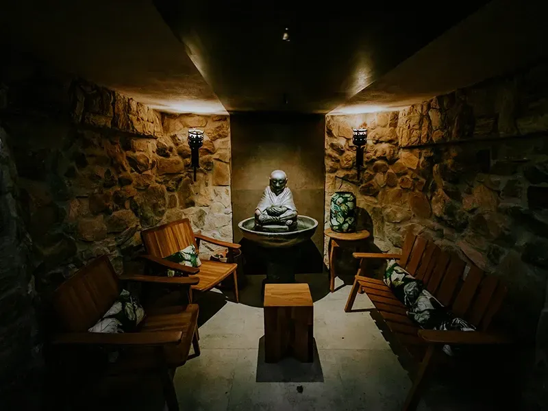 Uma sala com paredes de pedra e iluminação tênue, com bancos de madeira voltados para uma estátua central em uma bacia.