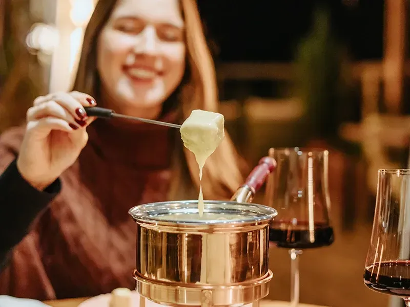 Uma pessoa sorri enquanto usa um garfo de fondue para mergulhar um pedaço de pão em uma panela de queijo derretido ao lado de taças de vinho.