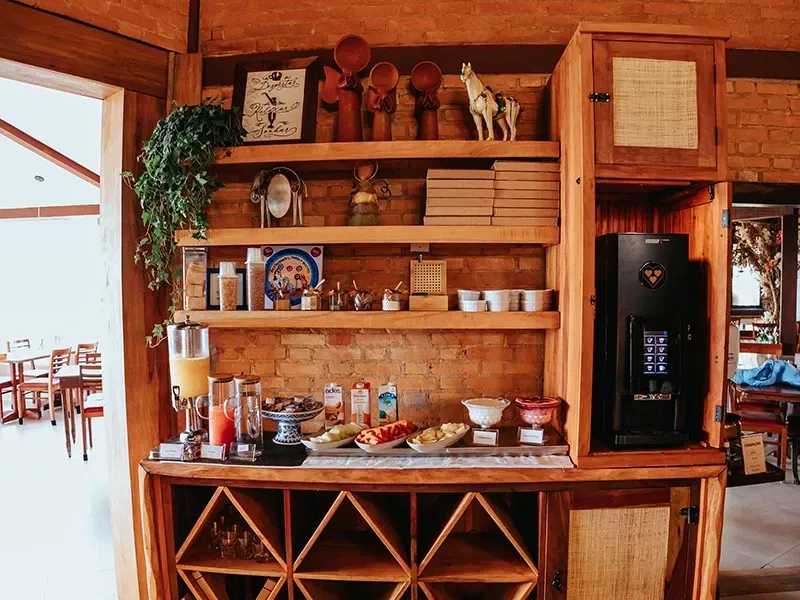 Estação de buffet de café da manhã de hotel com prateleiras de madeira contendo lanches, bebidas e uma máquina de café encostada em uma parede de tijolos.