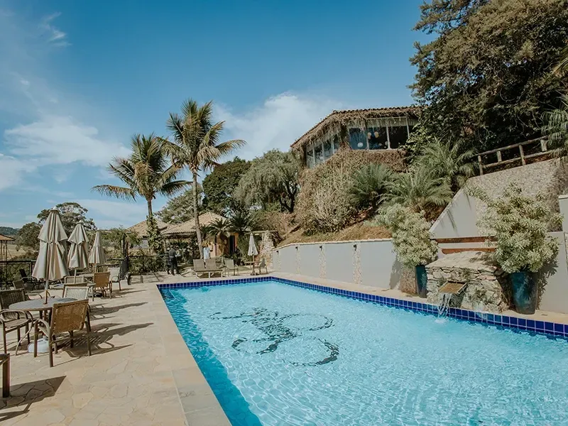 Uma piscina de resort com um mosaico no fundo, rodeada por espreguiçadeiras, palmeiras e um edifício na encosta de uma colina.