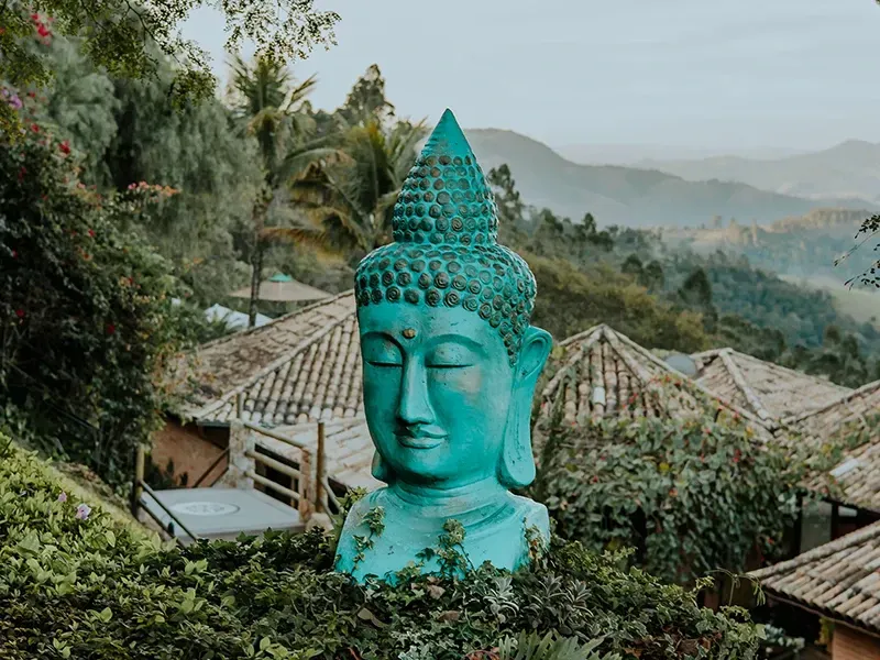 Uma escultura de uma cabeça de Buda em tons de turquesa se destaca em meio a uma vegetação exuberante, com telhados de telha e colinas montanhosas envoltas em névoa ao fundo.