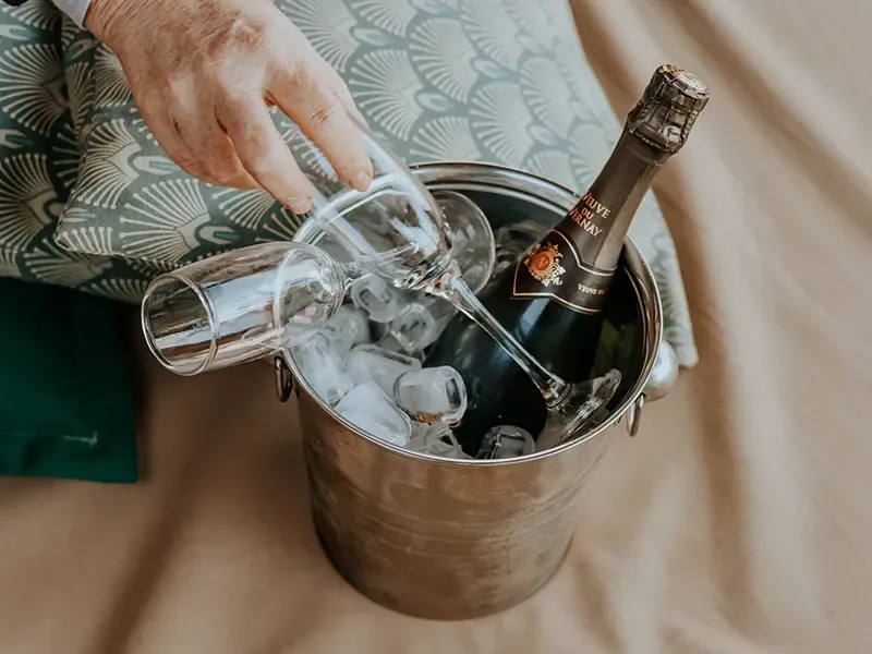 Uma mão se estende em direção a uma taça de champanhe que repousa em um balde de gelo de metal contendo uma garrafa de vinho espumante.