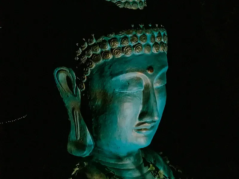 Uma estátua em tons turquesa da cabeça de Buda em perfil contra um fundo escuro, realçada por uma iluminação suave e direcionada.