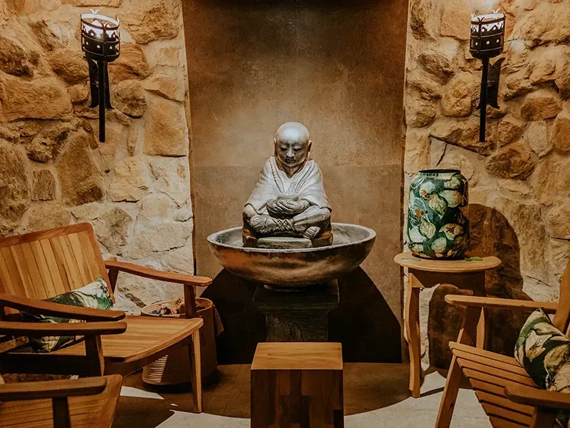 Um recanto sereno de spa com duas cadeiras de madeira, uma estátua de Buda de pedra como elemento central e uma fonte de água, além de iluminação aconchegante nas paredes de pedra.