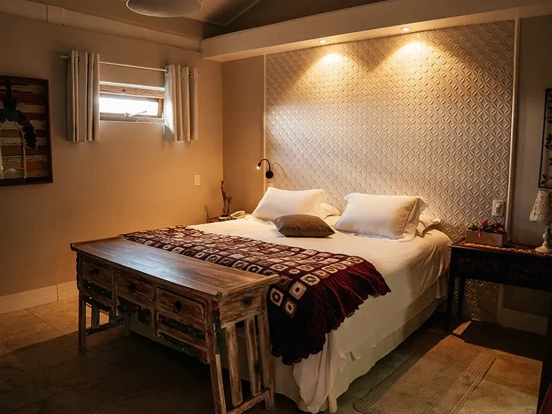 Um quarto com uma cama de casal, uma cabeceira estampada, um banco de madeira aos pés da cama e iluminação aconchegante no teto.