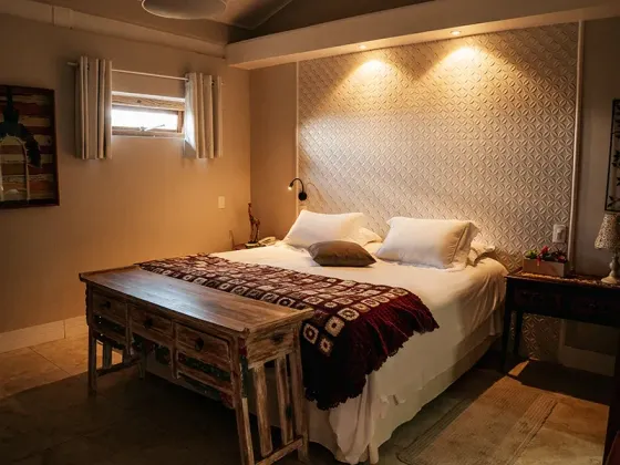 Um quarto com uma cama de casal, uma cabeceira estampada, um banco de madeira aos pés da cama e iluminação aconchegante no teto.