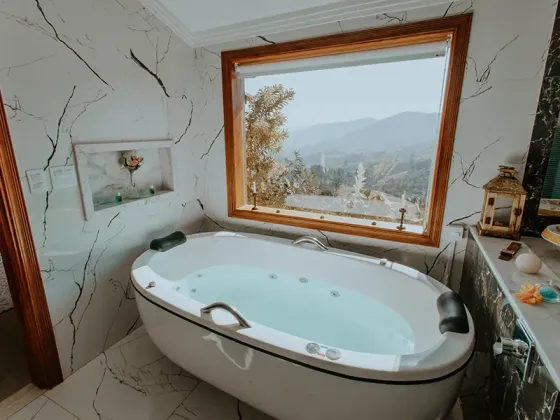 Uma banheira de hidromassagem branca em um banheiro revestido de mármore, com uma grande janela que oferece uma vista panorâmica das montanhas e árvores.