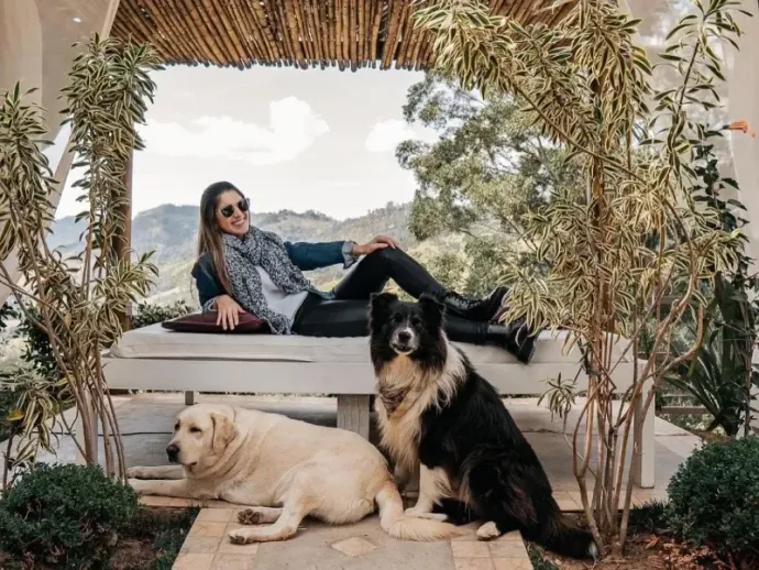 Uma pessoa de óculos escuros está sentada em uma espreguiçadeira ao ar livre, com um labrador amarelo e um cachorro preto e branco descansando por perto.