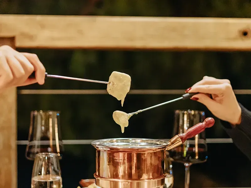 Duas pessoas mergulham alimentos em uma panela de cobre para fondue de queijo, em uma mesa de madeira com taças de vinho por perto.
