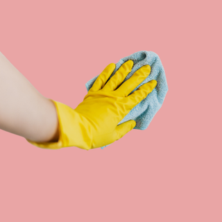 Yellow-gloved hand holding a blue cleaning cloth on a pink background.