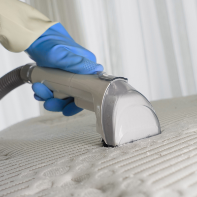 Person wearing blue gloves cleaning a white mattress with a handheld cleaning tool.
