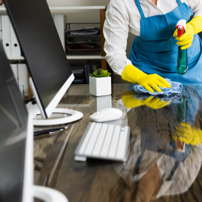 Person in blue apron and yellow gloves cleaning an office desk with spray bottle and cloth.