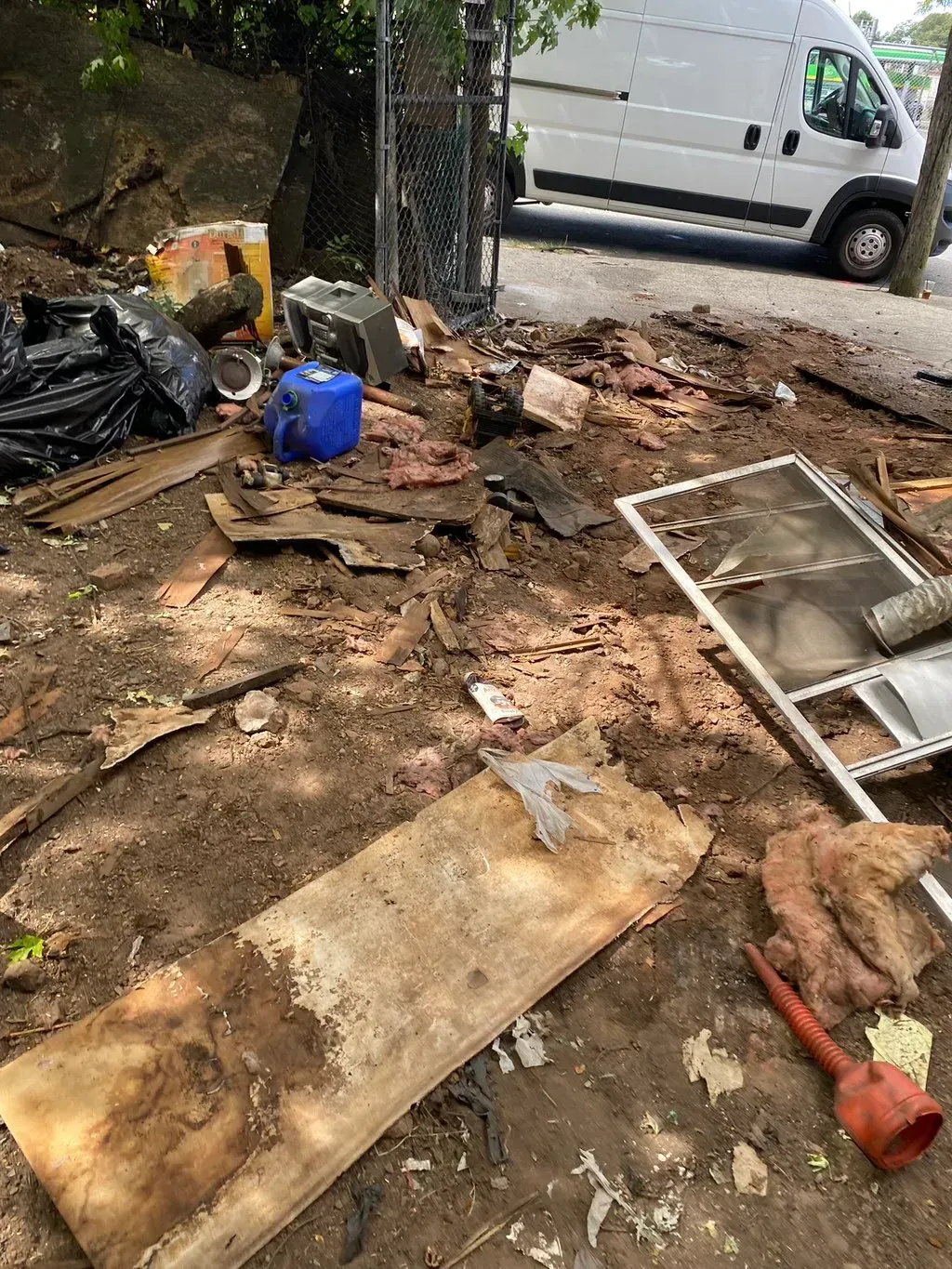 Garbage pile on dirt and concrete. Includes wood, plastic, and a blue container near a fence and van.