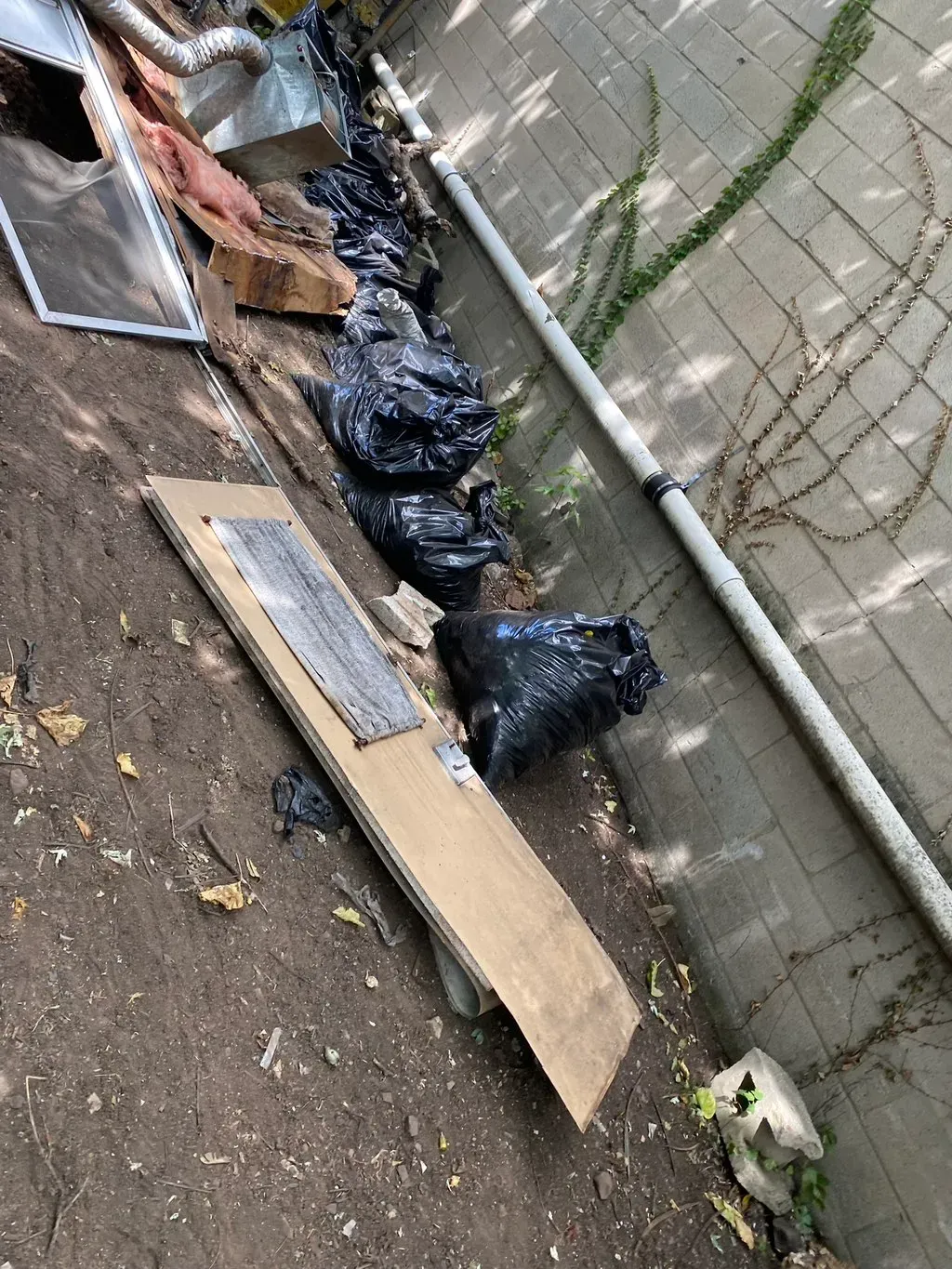 Debris, trash bags, and a board along a wall and dirt. Outdoors.