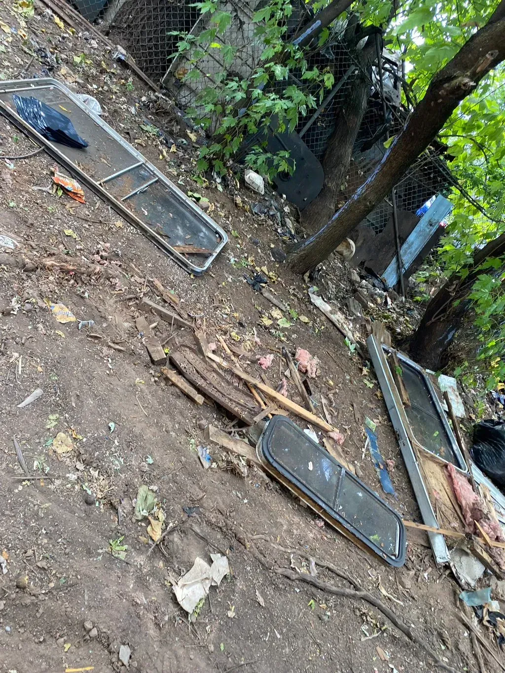 Debris, including window frames, litter a muddy hillside next to a tree and fence.