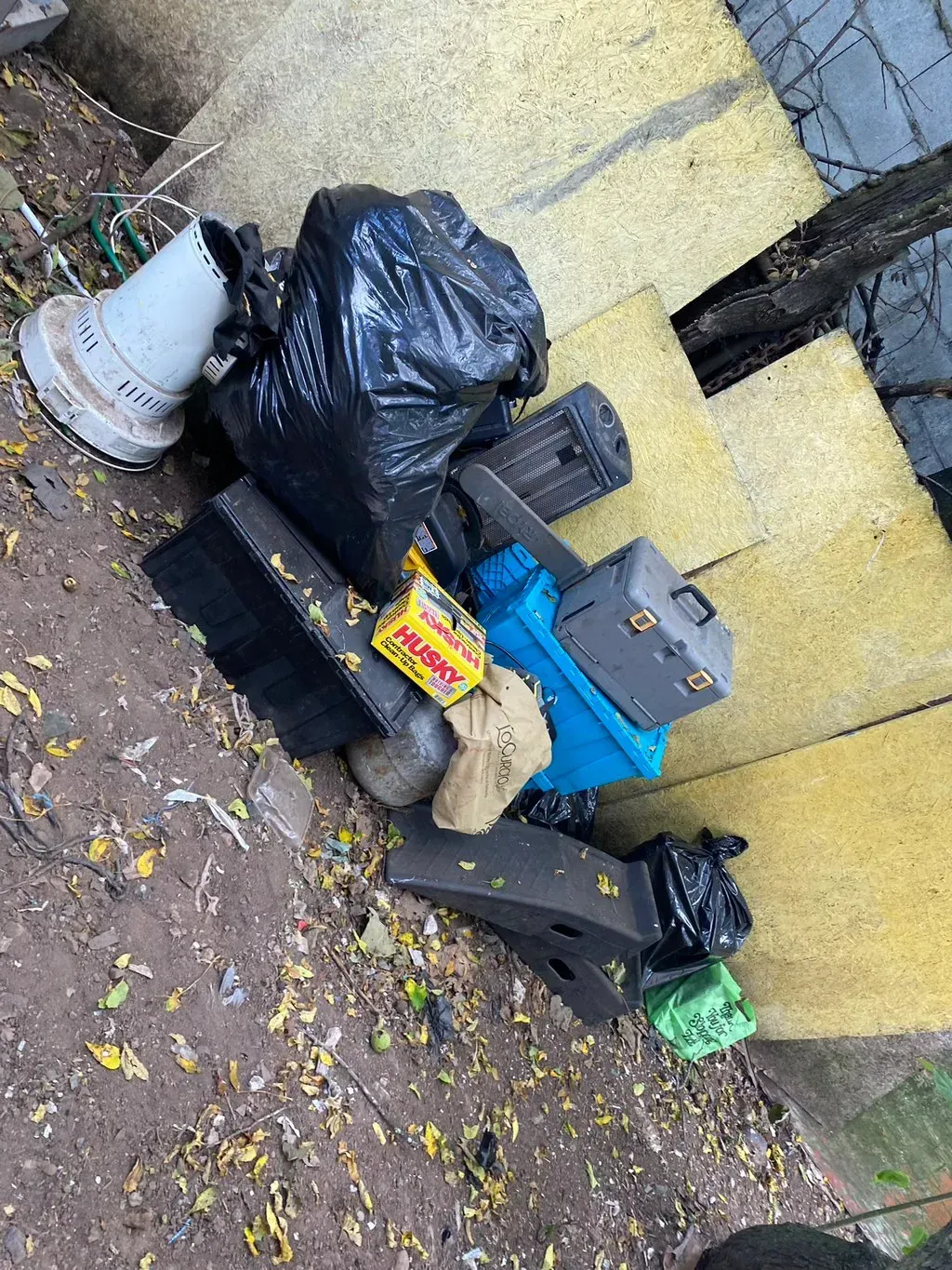 Pile of trash including bags, boxes, and a bird feeder on the ground next to a yellow-toned wall.