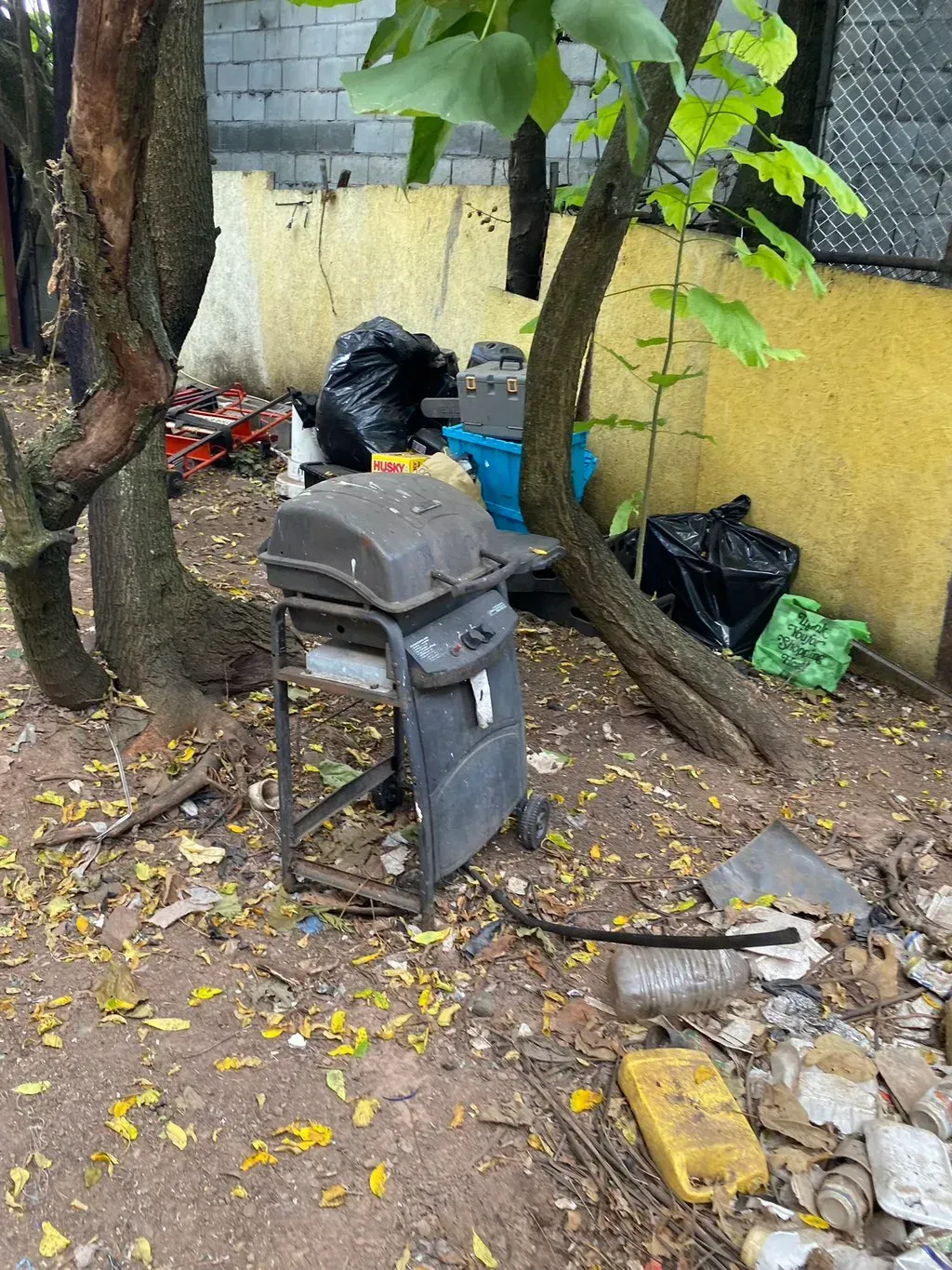 Grill surrounded by trash, fallen leaves, and two trees in a yard.