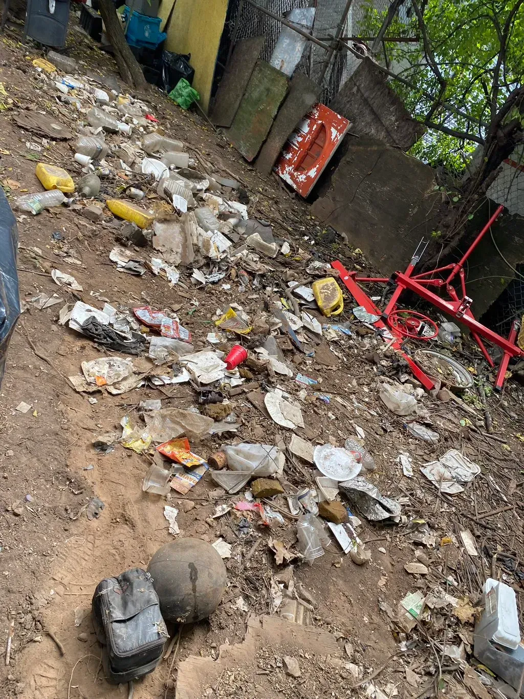 Debris-strewn hillside: trash, plastic, and a red object lie scattered on dirt and rocks outdoors.
